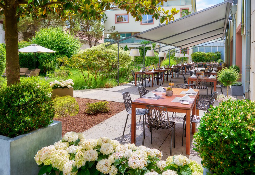 Sommer-Terrasse des Restaurants RIVA im Weinhotel Kaisergarten Deidesheim/ Pfalz. Die großzügige Terrasse wird von einer sandfarbenen Markise beschattet. Die Tische sind einladend eingedeckt. Um die Terrasse herum ist der mediterran angelegte Garten zu sehen.